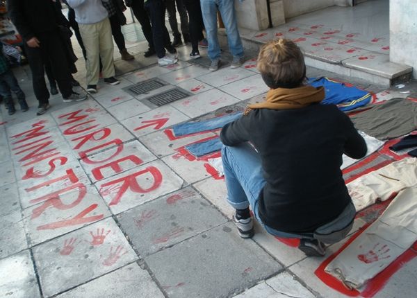 Φωτογραφία: Στην είσοδο του Υπουργείο νεαρή διαδηλώτρια βάφει κόκκινο το περίγραμμα αντρικών ρούχων. Κόκκινη μπογιά γράφει στο πεζοδρόμιο με κεφαλάια Murder Ministry και ένα βελάκι δείχνει την είσοδο. Κόκκινα αποτυπώματα χεριών παντού.