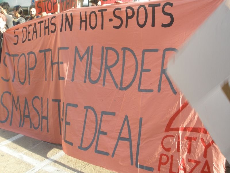 Φωτογραφία: Πορτοκαλί πανό γράφει με κεφαλαία: 5 DEATHS IN HOT SPOTS. STOP THE MURDER. SMASH THE DEAL. Υπογράφεται από  ένα τριγωνάκι (στέγη) και "city plaza"
