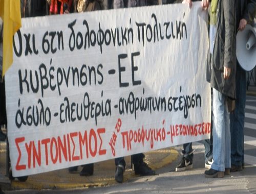Φωτογραφία: Άσπρο πανό γράφει με μάυρα γράμματα: Όχι στη δολοφονική επίθεση κυβέρνησης- ΕΕ. άσυλο- ελευθερία- ανθρώπινη στέγαση. Υπογραφή με κόκκινα γράμματα: ΣΥΝΤΟΝΙΣΜΟΣ για το προσφυγικό- μεταναστευτικό